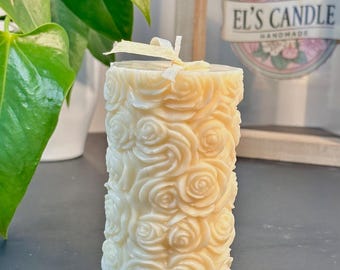 Può includere: Candela color avorio con motivo a rose. La candela cilindrica è adornata con rose in rilievo e legata con un nastro color crema. La candela è alta circa 10 cm. Lo sfondo include una pianta verde e un cartello con la scritta "EL'S CANDLE HANDMADE".