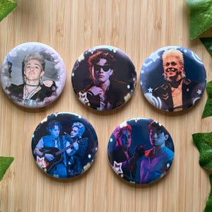 Puede incluir: Cinco chapas redondas con retratos fotográficos de dos personas. Los diseños incluyen tomas individuales e imágenes en dúo con una guitarra, acentuadas por gráficos de estrellas y corazones blancos sobre fondos oscuros, azules y morados.