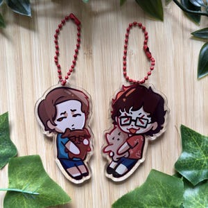 May include: Two clear acrylic keychains featuring cartoon-style characters. The left character has brown hair and a blue shirt, holding a brown creature. The right character has dark hair, glasses, and a red patterned shirt, holding a tan creature.