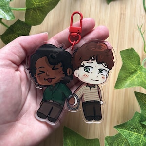 May include: A clear acrylic keychain featuring two cartoon characters connected by a small metal chain. One character has dark hair and a green shirt, while the other has curly brown hair and a striped shirt. A red lobster clasp is attached at the top.