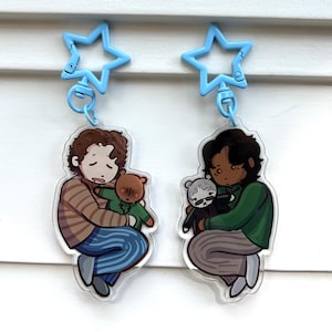 May include: Two acrylic keychains featuring illustrated characters with light blue star-shaped clasps. One character has curly brown hair, a striped shirt, and blue pants, holding a small bear. The other has dark hair, a green shirt, and gray pants.