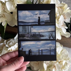 May include: A rectangular art print featuring three horizontal panels depicting coastal scenes. Each panel shows a blue sky with clouds, a body of water, and silhouettes of figures near buildings or fences. The overall color palette is shades of blue, white, and black.