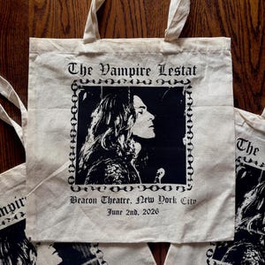 Op de afbeelding: Crèmekleurige draagtas met een zwart-wit portret en tekst. De tekst luidt "The Vampire Lestat", "Beacon Theatre, New York City, June 2nd, 2026". De afbeelding is omlijst met een kettingontwerp.