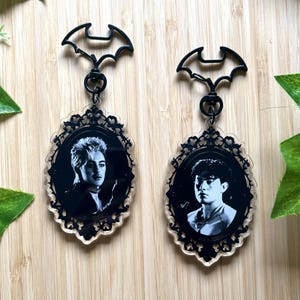 Peut inclure: Deux porte-clés en acrylique avec des fermoirs noirs en forme de chauve-souris et des cadres ovales aux bords en filigrane ornementés. Chaque pendentif présente un portrait en noir et blanc, l'un avec une coiffure hérissée, l'autre aux cheveux courts.