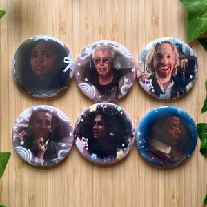 May include: A set of six circular pin-back buttons featuring photographic portraits of various individuals. Each button is decorated with a white or colorful graphic overlay of stars, swirls, and ribbons against the central character image.