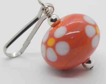 May include: A small, orange glass bead with white and yellow flower designs. The bead is attached to a silver metal clasp.