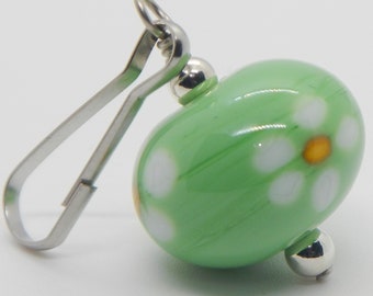 May include: A green glass bead with white daisy designs and a yellow center. The bead is attached to a silver metal clip.