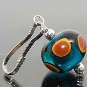 Peut inclure: Une perle en verre bleu turquoise avec des accents orange et noirs pend d'une boucle en métal argenté. La perle a une finition brillante et lustrée.
