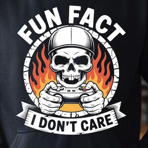 May include: Black hoodie featuring a graphic of a skull wearing a cap and holding a game controller, surrounded by flames. The text reads "FUN FACT I DON'T CARE" in white. The hoodie has a drawstring hood and a kangaroo pocket.