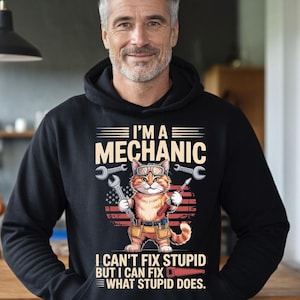 Könnte beinhalten: Schwarzer Kapuzenpullover mit einem Cartoon-Mechaniker-Kater, der Werkzeuge hält. Der Text lautet "I'M A MECHANIC" und "I CAN'T FIX STUPID BUT I CAN FIX WHAT STUPID DOES". Die Katze trägt eine Brille und einen Werkzeuggürtel.