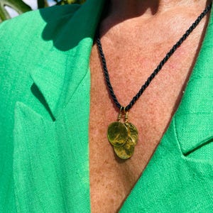 Op de afbeelding: Een goudkleurige hangerketting met drie cirkelvormige bedels, hangend aan een zwart koord. De ketting wordt getoond tegen een groene jas.