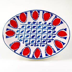 Może przedstawiać: Owalny ceramiczny półmisek z białym tłem i centralnym niebieskim wzorem w kratkę. Brzeg ozdobiony jest powtarzającym się motywem czerwonych elementów w kształcie migdałów, otoczonych małymi niebieskimi kropkami i cienką niebieską obwódką.