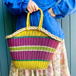 May include: A woven basket bag featuring horizontal stripes in yellow, purple, and lime green. The bag has a curved top edge with a braided natural-colored rim and two yellow circular handles. The structure is made of thin, flexible material.