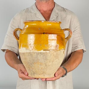 Op de afbeelding: Een grote keramische pot met twee oren, voorzien van een glanzend honinggeel geglazuurd bovengedeelte en een matte, ongeglazuurde beige onderkant. De pot heeft een brede, ronde opening en een bolvormig lichaam, met een duidelijk contrast tussen de gladde, glanzende bovenkant en de verweerde, textuurrijke onderzijde.