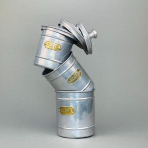 May include: Three silver metal storage canisters, stacked with lids. Each canister has a gold label, reading "PATES", "CAFE", and "SUCRE". The top lid is partially open, revealing the contents.