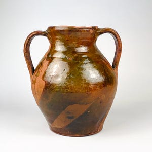 May include: A rustic earthenware vessel featuring two curved handles and a wide mouth. The exterior displays a glossy, mottled glaze in shades of deep brown, ochre, and dark green, with patches of unglazed reddish-brown clay visible on the surface.