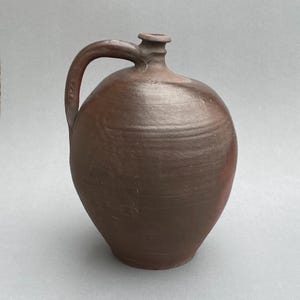 May include: A brown ceramic jug with a rounded body and a single handle. The jug has a slightly flared opening at the top.
