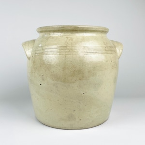 May include: A beige ceramic crock with a wide mouth and two side handles. The exterior features a smooth, glazed finish with subtle speckling and a series of horizontal incised lines encircling the upper portion of the vessel below the rim.