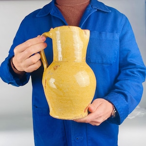 Puede incluir: Una jarra de cerámica amarilla con asa y pico. La jarra tiene un cuerpo redondeado y una superficie texturizada. La jarra está sostenida por una persona que lleva una chaqueta azul.