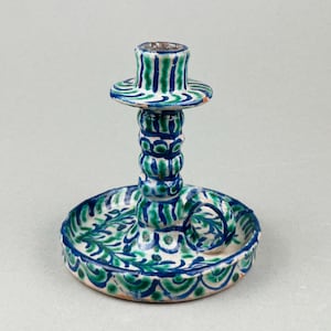 Può includere: Un candelabro in ceramica bianca con un motivo floreale verde e blu. Il candelabro ha una base rotonda e uno stelo alto e sottile con una coppa per tenere una candela.