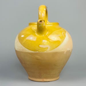 May include: A yellow and tan ceramic pitcher with a handle and spout. The pitcher has a rounded body and a handle that arches over the top. The spout is located on the front of the pitcher. The pitcher is likely vintage or antique.