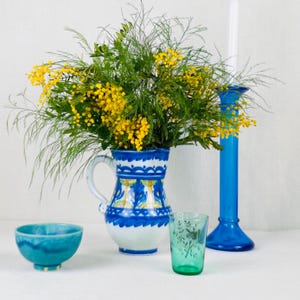 Puede incluir: Naturaleza muerta con una jarra de cerámica blanca y azul con flores amarillas, un cuenco turquesa, un vaso verde y un candelabro azul con una vela blanca. Los objetos están dispuestos sobre una superficie blanca.