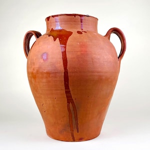 May include: A large terracotta vessel with two side handles and a wide mouth. The surface is a warm reddish-brown clay color with a glossy, dark brown glaze drip pattern running vertically from the rim down the front of the rounded body.