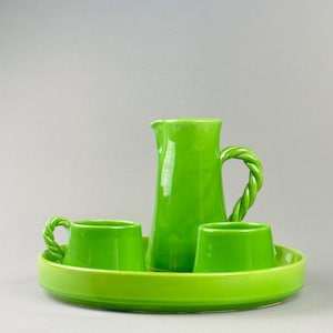 Puede incluir: Un juego de cerámica verde brillante que incluye una jarra con asa trenzada, dos tazas a juego y una bandeja redonda. El conjunto es de un color verde vibrante y sólido, perfecto para servir bebidas. La jarra mide unos 20 cm de alto.