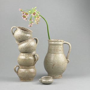 Puede incluir: Un juego de cuatro tazas de cerámica apiladas con un esmalte moteado, junto a una jarra de cerámica más grande con un esmalte similar. Un pequeño cuenco de cerámica se encuentra delante de las tazas. Una rama de flores rosas se encuentra en el fondo.