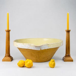 May include: A large, round, tan ceramic mixing bowl with a wide, cream-colored rim and a small pouring spout. The bowl is flanked by two tall, fluted wooden candle holders, each topped with a yellow taper candle. Three yellow quinces sit in front.