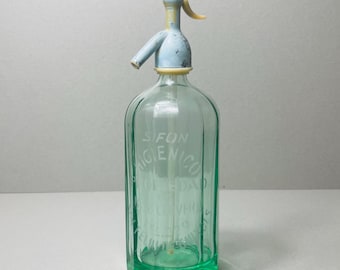 Pode incluir: Garrafa de sifão de vidro vintage com um dispensador azul claro e amarelo. A garrafa tem uma tonalidade verde e o texto "SIFON HIGIENICO" é visível. Uma peça clássica para refrigerantes ou água.