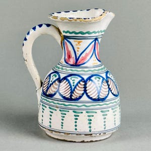 Puede incluir: Jarra pequeña de cerámica blanca con asa y pico. Decorada con patrones geométricos azules, verdes, rosas y marrones. El borde está adornado con pintura dorada. La jarra mide aproximadamente 15 cm de altura.