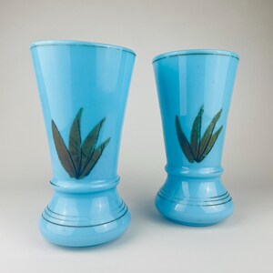 May include: Two light blue glass vases with a flared top and a decorative base. Each vase features a painted design of green and brown leaves. The rims are accented with a thin gold line.