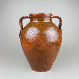 Peut inclure: Un vase en céramique marron avec deux anses. Le vase a un corps arrondi et un bord légèrement évasé. Des lignes ondulées décoratives encerclent la partie supérieure du vase. La couleur est un mélange de marron et de brun rougeâtre.