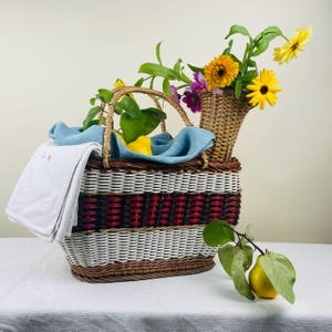 Könnte beinhalten: Ein Picknickkorb aus Weide mit Griff, mit einem rot-weißen Muster. Der Korb ist mit einem blauen Tuch, Zitronen und einem weißen Handtuch gefüllt. Eine kleine gewebte Vase enthält gelbe und lila Blumen. Eine gelbe Frucht und ein Zweig mit grünen Blättern liegen daneben.