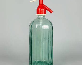 May include: Vintage glass seltzer bottle with a red and white dispenser top. The bottle is a clear, light green colour with vertical ridges. The dispenser has a lever and a spout.
