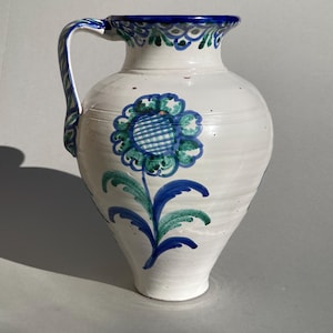 Op de afbeelding: Witte keramische kan met een blauw en groen bloemmotief. De kan heeft een gebogen handvat en een brede opening met blauwe rand. Het bloemmotief heeft een grote blauw-groene bloem met een geblokt centrum en blauwe bladeren.
