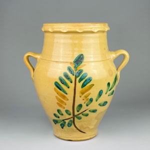 May include: A yellow ceramic vase with two handles and a scalloped rim. The vase is decorated with a painted leaf design in shades of blue, green, and yellow. The vase has a rounded body and a slightly flared top.
