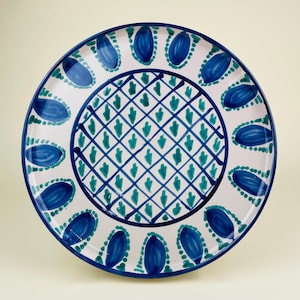 Può includere: Un piatto in ceramica bianca con un design geometrico blu e turchese. Il piatto presenta un motivo a griglia centrale e forme ovali attorno al bordo, il tutto in tonalità di blu e turchese. Il piatto ha un bordo blu.