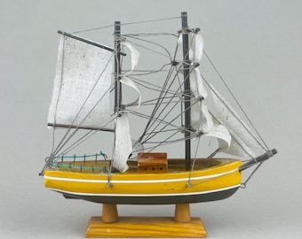 May include: A miniature wooden sailboat model. The hull is painted yellow with a black base and white trim. The sails are white, and the rigging is black. The model sits on a wooden stand, and the overall design is nautical.