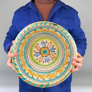 May include: A hand-painted ceramic plate with a floral design. The plate features a central flower in blue, orange, and green, surrounded by concentric rings of yellow, green, and brown patterns. The plate is held against a blue background.