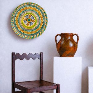May include: A round ceramic plate with a hand-painted floral pattern in yellow, green, and blue, mounted on a wall. Beside it sits a dark brown glazed ceramic jug with two handles, positioned on a white pedestal next to a dark wooden chair.