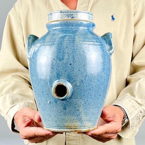 Op de afbeelding: Een lichtblauwe keramische pot met een gespikkelde afwerking, voorzien van twee zijhandvatten, een cilindrische hals en een ronde opening aan de onderzijde. Het vat heeft een gladde, geglazuurde textuur met subtiele horizontale ribbels.