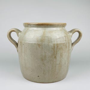 May include: A vintage stoneware crock with two curved handles. The off-white ceramic pot has a slightly flared rim and horizontal banded detailing. The exterior features subtle brown streaks and a light brown rim.