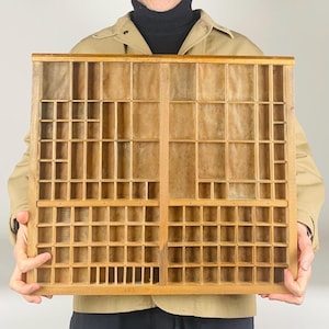 May include: A vintage wooden printer's tray with numerous small compartments of varying sizes. The tray is rectangular, with a light brown wood finish and a grid-like structure. The compartments are designed to hold individual letters or other small items. The tray is held by a person.