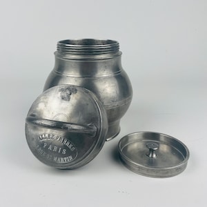 May include: A vintage metal container with a lid and a small, separate lid. The main lid has a handle and the text "ALLEZ FRERES PARIS 7 RUE ST MARTIN". The container is made of a dull, silver-colored metal.