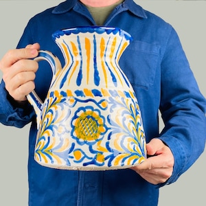 Può includere: Una grande brocca in ceramica dipinta a mano, con bocca larga e manico. La brocca presenta una base bianca con disegni floreali e a righe blu e gialli. Il design include un motivo a girasole.