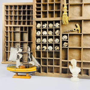 May include: A vintage wooden printer's tray with numerous compartments, containing speckled eggs. A small yellow sailboat with white sails and a bust statue are in front. A gold tassel and corkscrew are also visible.