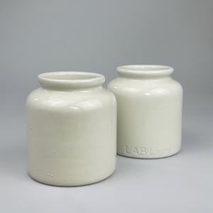 Op de afbeelding: Twee crèmekleurige keramische potten met afgeronde lichamen en licht uitlopende randen. Eén pot heeft de tekst "LAB Lagny" in reliëf op de zijkant. De potten staan tegen een effen grijze achtergrond en tonen hun eenvoudige, klassieke ontwerp.