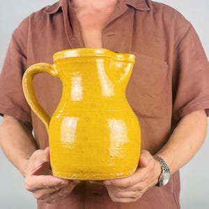 May include: A mustard yellow ceramic pitcher with a glossy finish, featuring a rounded body, a wide pouring spout, and a curved handle on the side. The surface shows subtle variations in color and texture, with a horizontal band around the middle.